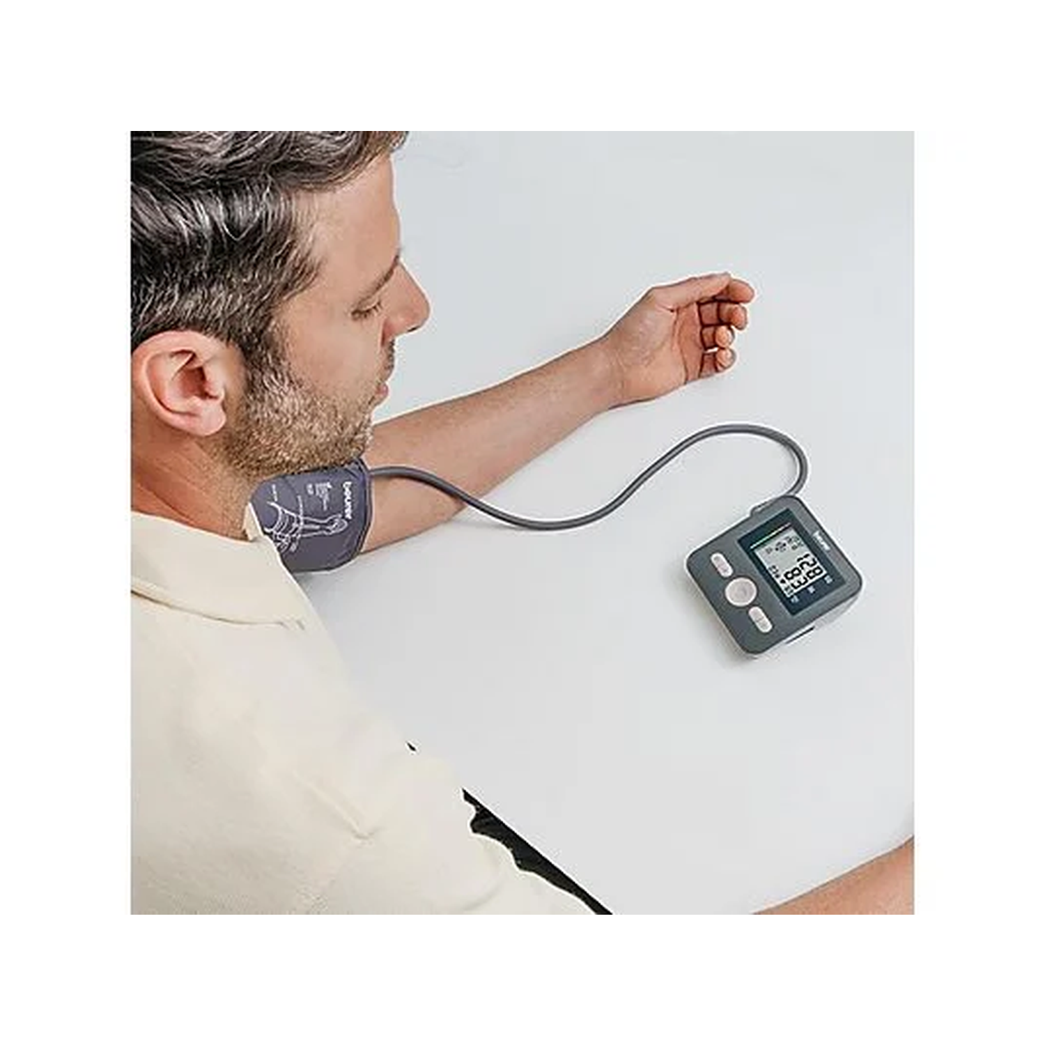 A man wearing a blood pressure monitor. The cuff is on his arm, and the device displays readings. He is on a white surface.