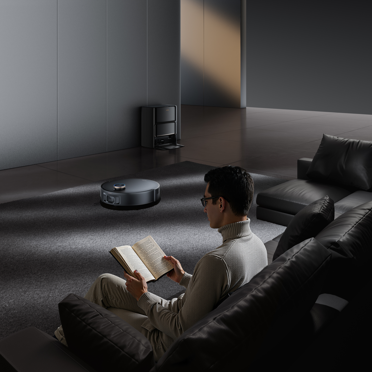 A man reads a book on a black leather sofa. A robotic vacuum cleaner is on the carpet. A charging station is behind.