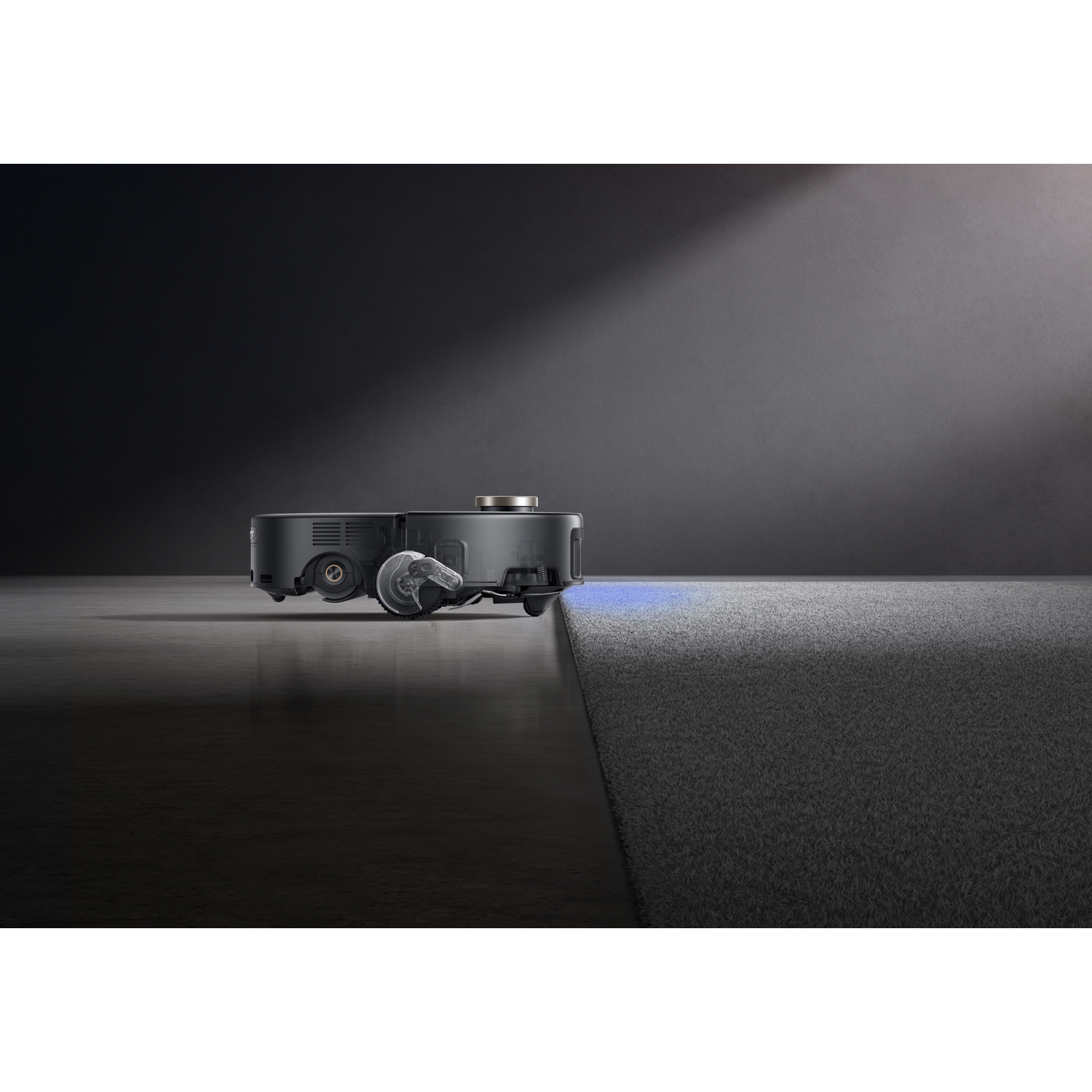 A black robot vacuum cleaner on a dark surface. It's positioned near a gray carpet. A light beam shines on the vacuum.