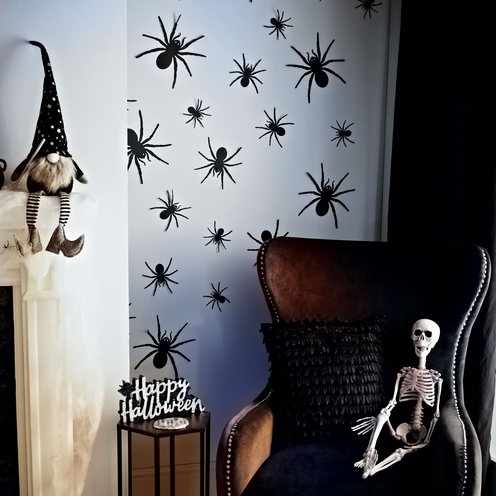 Halloween dekoráció: gnóm a kandallón, pókok a falon, csontváz a széken és Happy Halloween felirat.