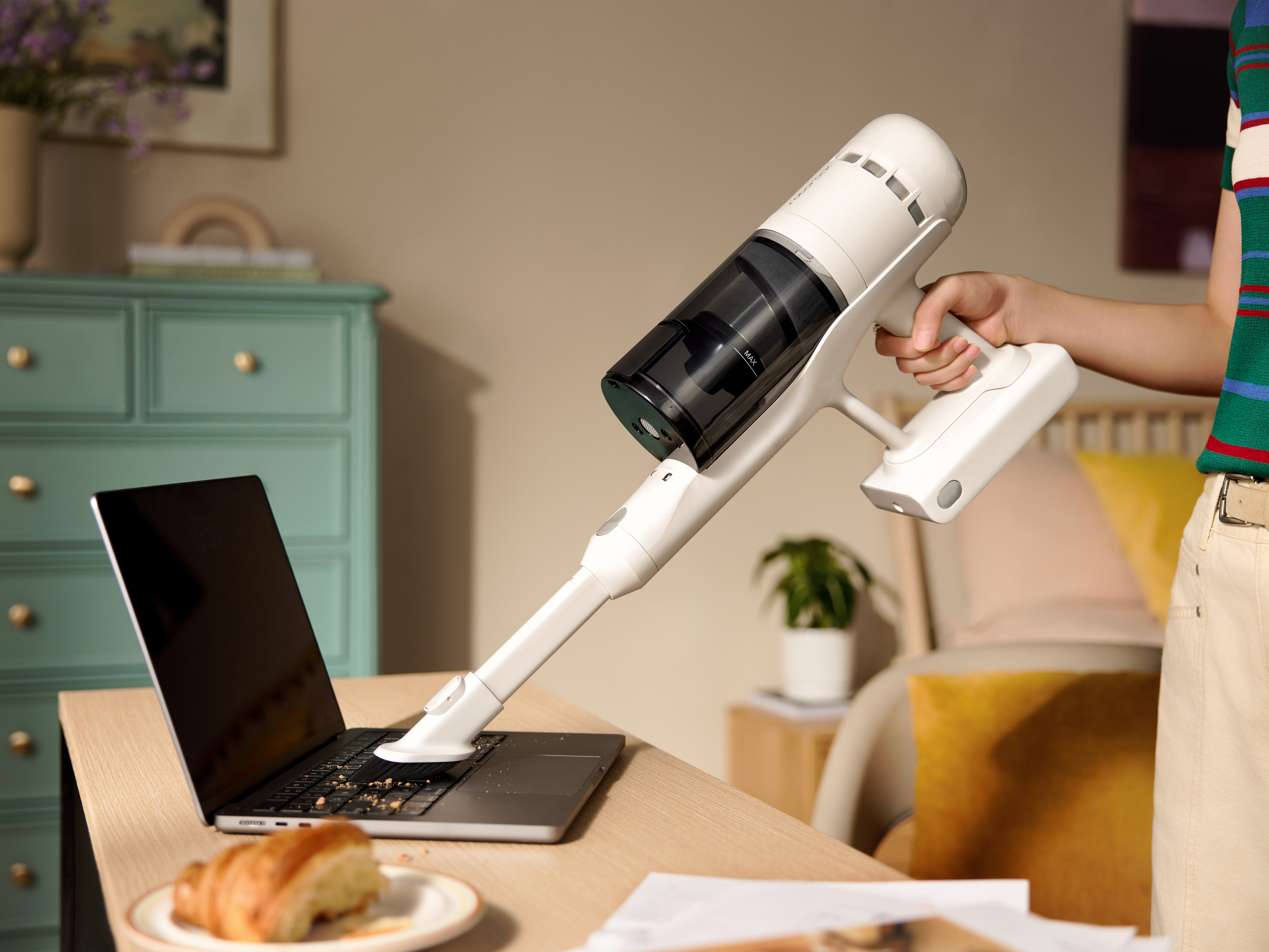 Eine Person, die eine Laptop-Tastatur mit einem weißen Staubsauger absaugt. Ein Croissant liegt auf einem Teller auf dem Schreibtisch.