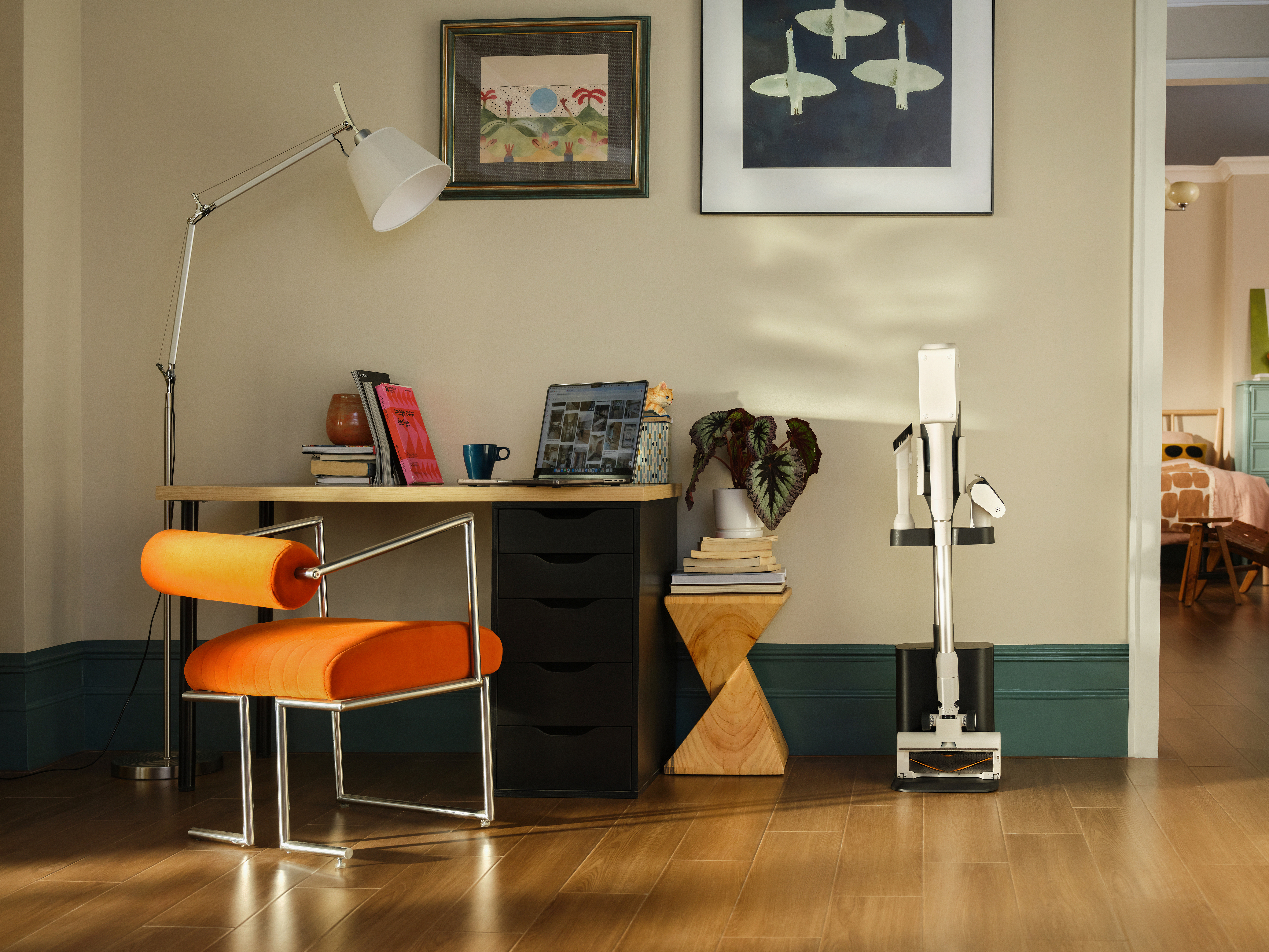 Ein Schreibtisch mit Laptop, Lampe und orangefarbenem Stuhl. Ein Staubsauger steht daneben. Kunst hängt an der Wand. Eine Tür führt zu einem Schlafzimmer.