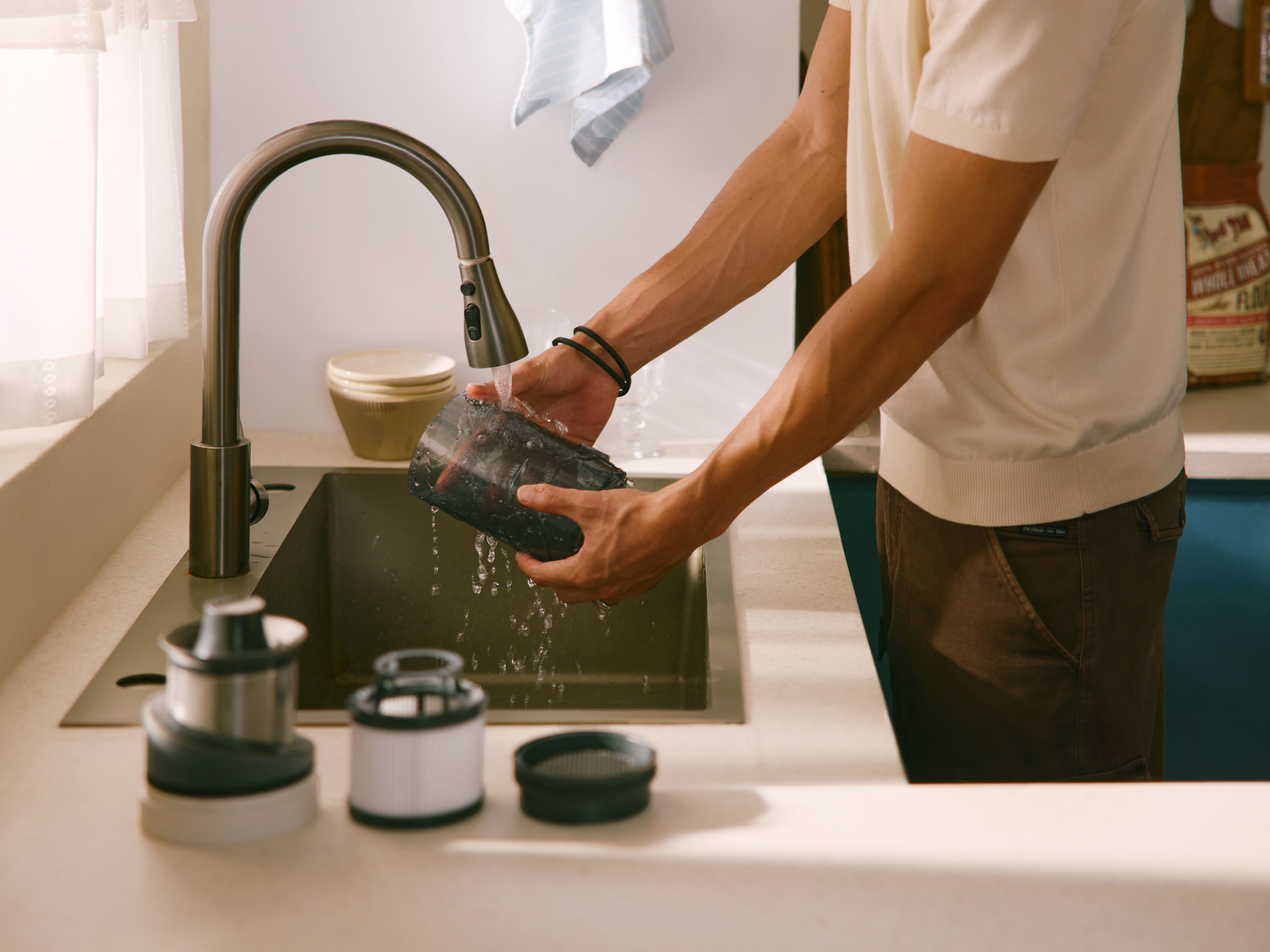 Eine Person wäscht ein Staubsaugerteil unter einem Küchenwasserhahn. Teile liegen auf der Theke. Helle Farben dominieren die Szene.