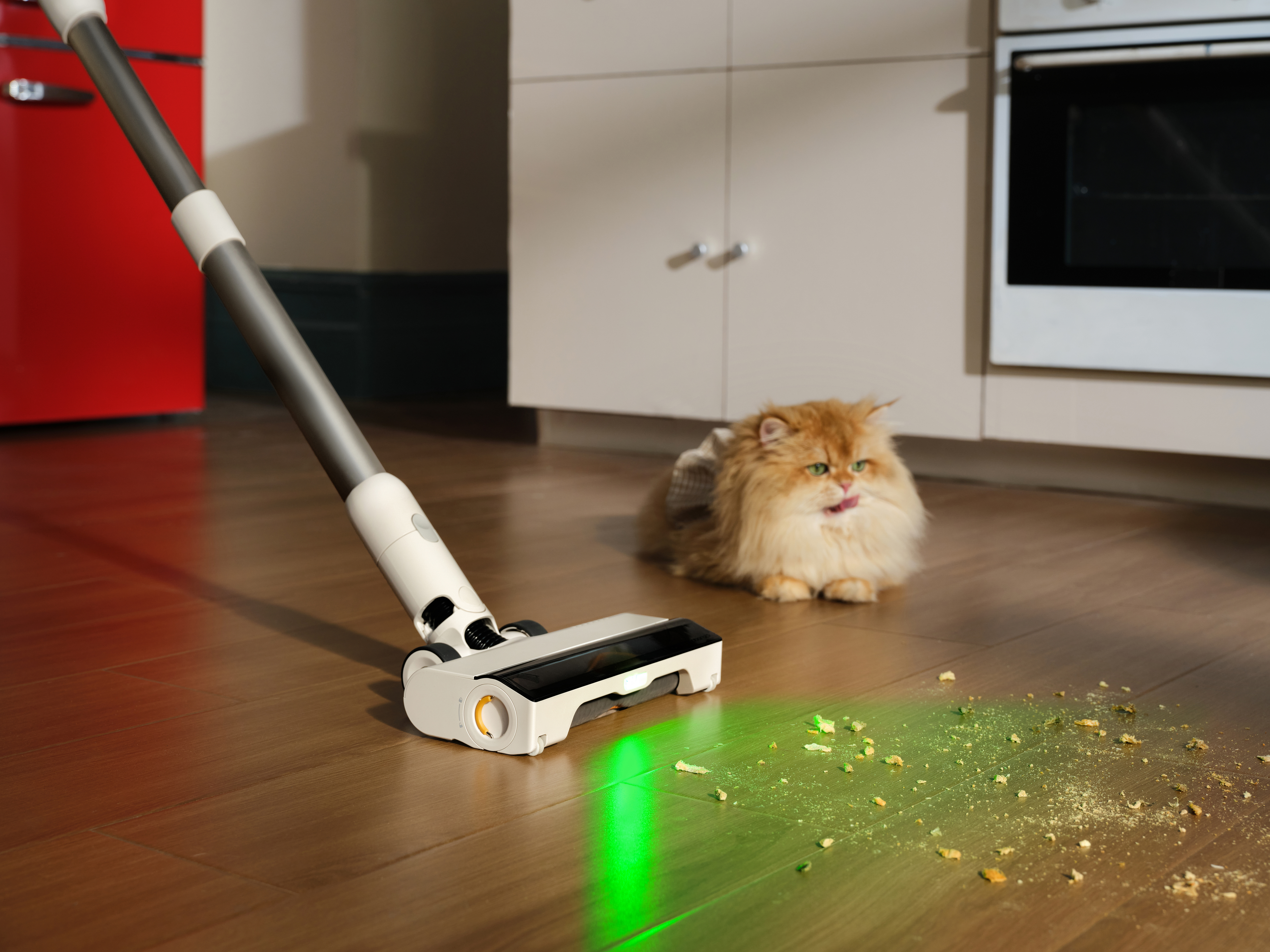 Ein Staubsauger reinigt Krümel auf einem Holzboden. Eine flauschige Katze sitzt in der Nähe. Im Hintergrund ist ein roter Kühlschrank zu sehen.
