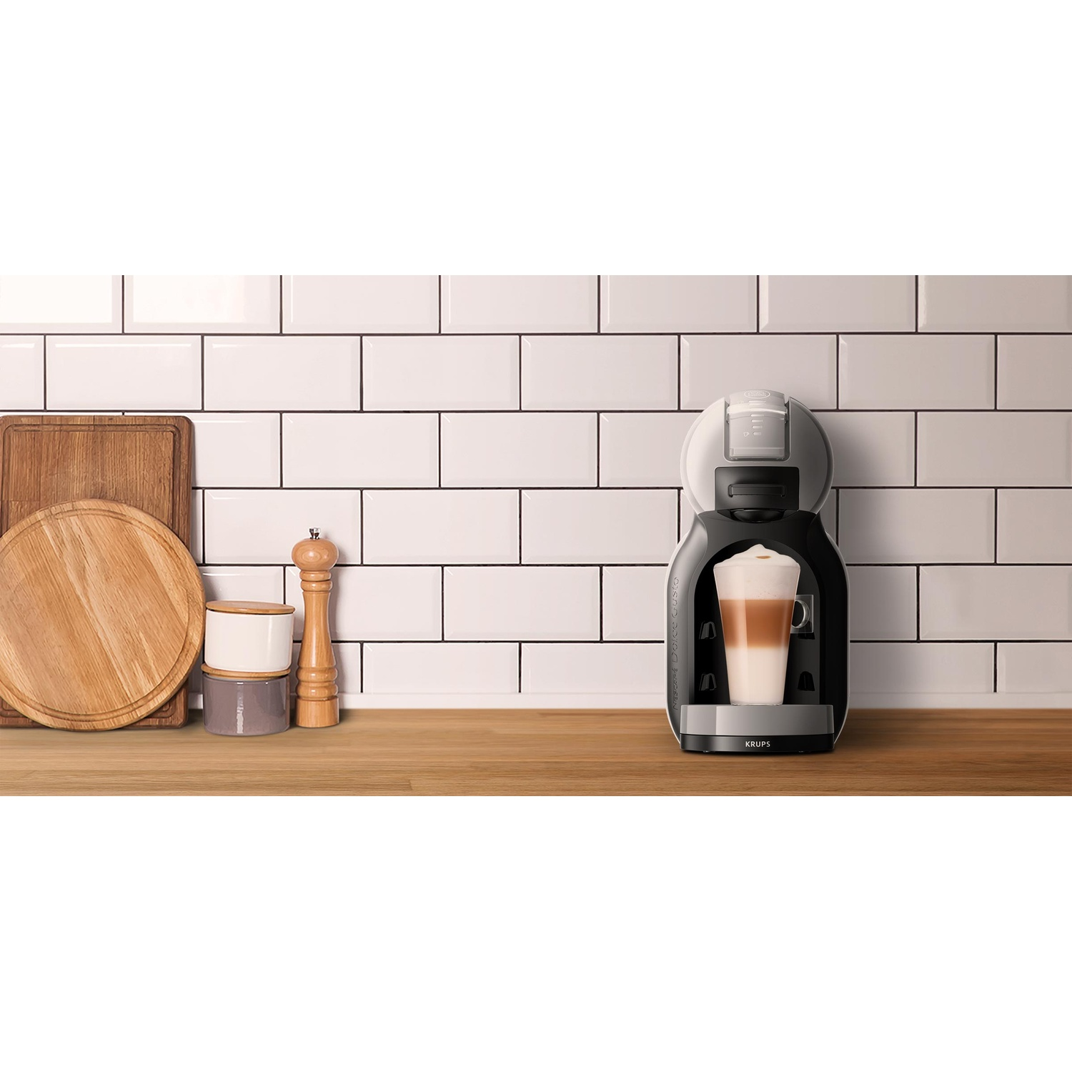 A coffee machine with a glass of layered coffee, cutting boards, a grinder, and jars, set against white tiles on a wooden surface.
