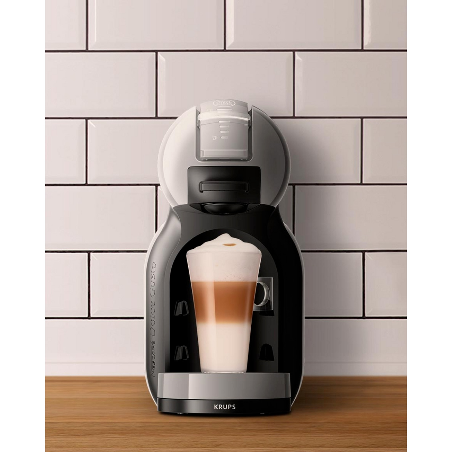 A black and white coffee machine with a glass of layered latte macchiato on a wooden table against white tiled wall.