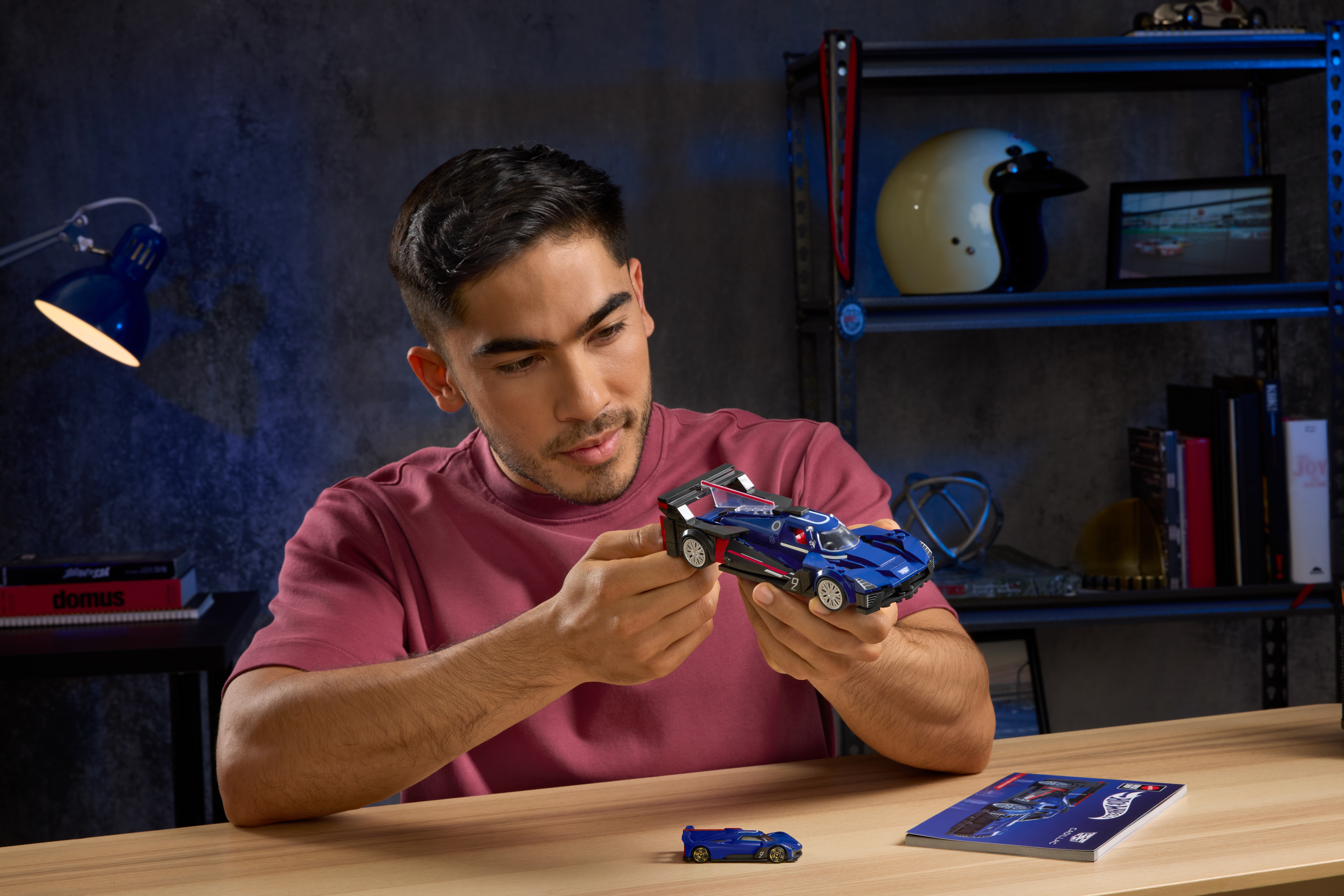 Ein Mann betrachtet ein blaues Spielzeug-Rennauto. Im Hintergrund sind ein kleineres Automodell, eine Zeitschrift und ein Helm zu sehen.