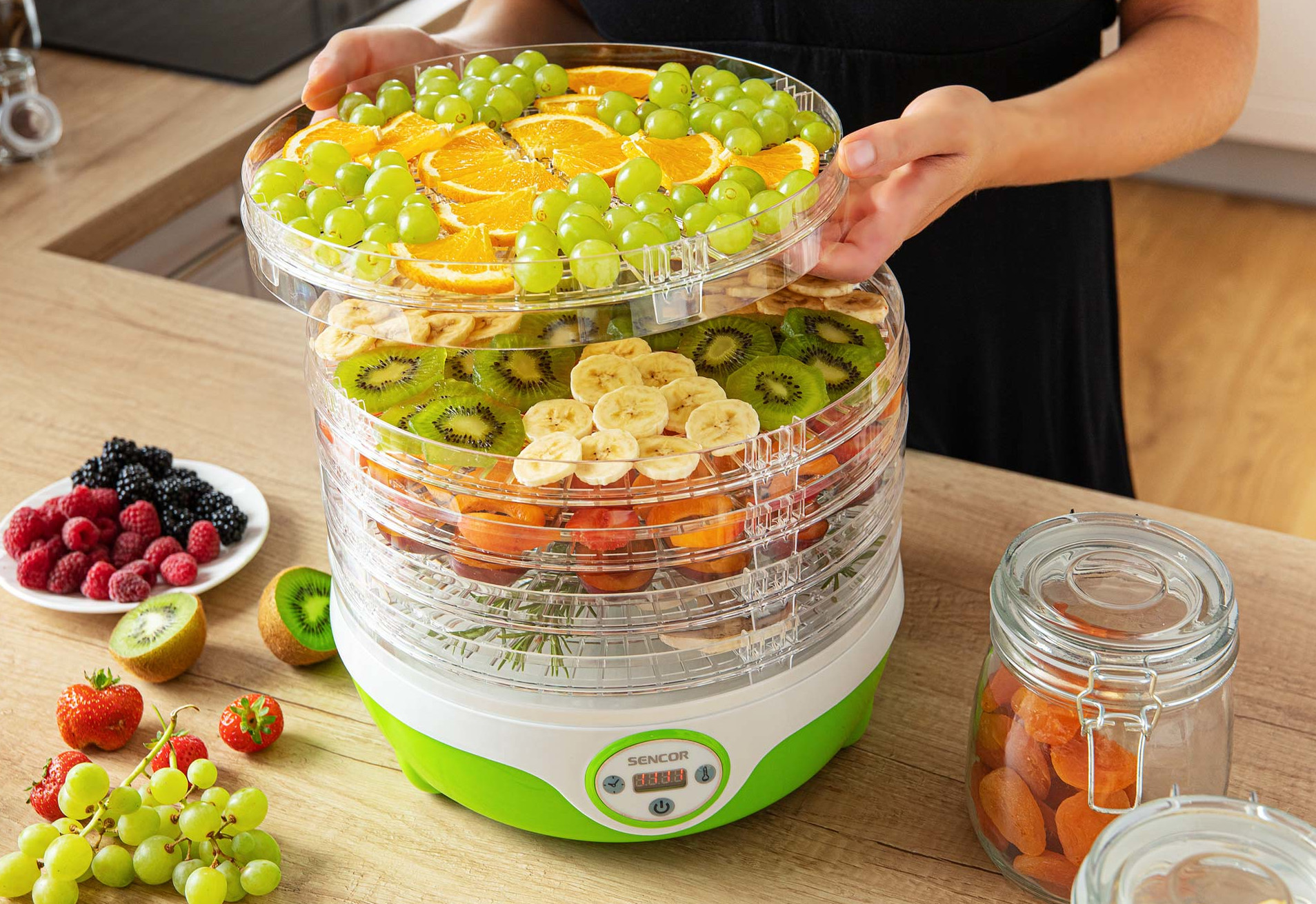 Osoba podnosi tacę z owocami z suszarki do żywności. Widoczne są winogrona, pomarańcze, kiwi i banany. Inne owoce otaczają suszarkę.