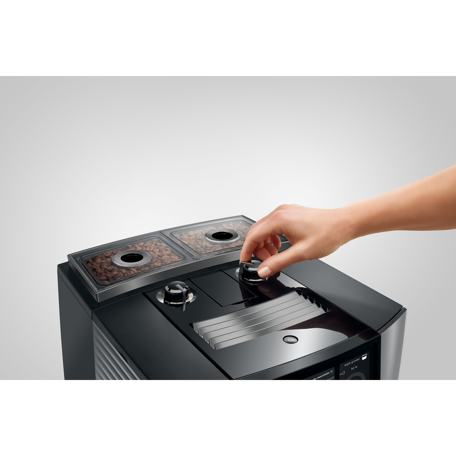 A hand turns a knob on a black coffee machine. Coffee beans are visible in transparent containers above. The background is white.