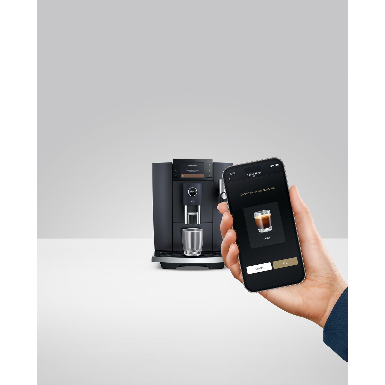 A black coffee machine with a glass and a phone showing coffee timer is held in hand.