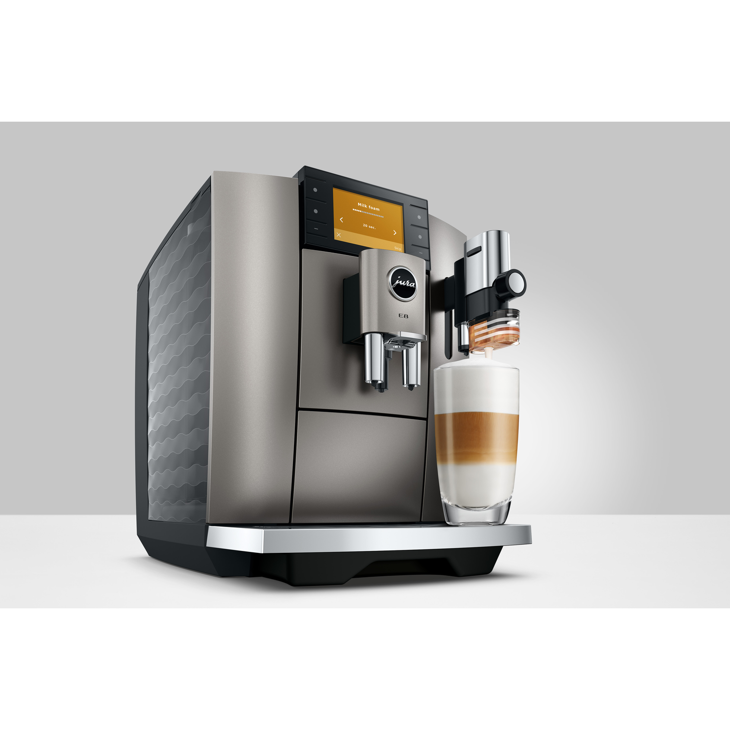 A Jura E8 coffee machine is making latte. A glass shows layers of milk, coffee, and foam. Machine is silver and black.
