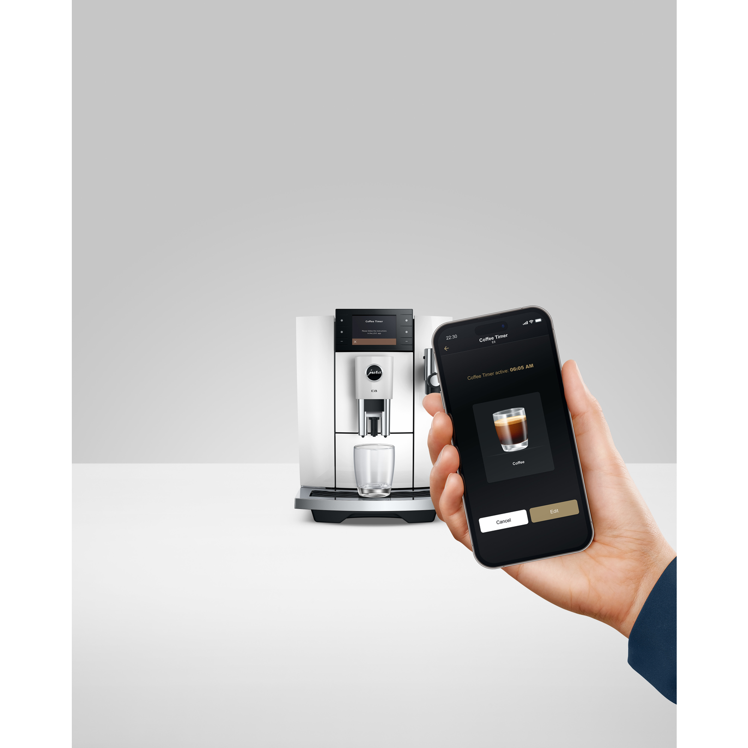 A white and silver coffee machine is next to a hand holding a phone displaying coffee timer. A glass is under the coffee spout.