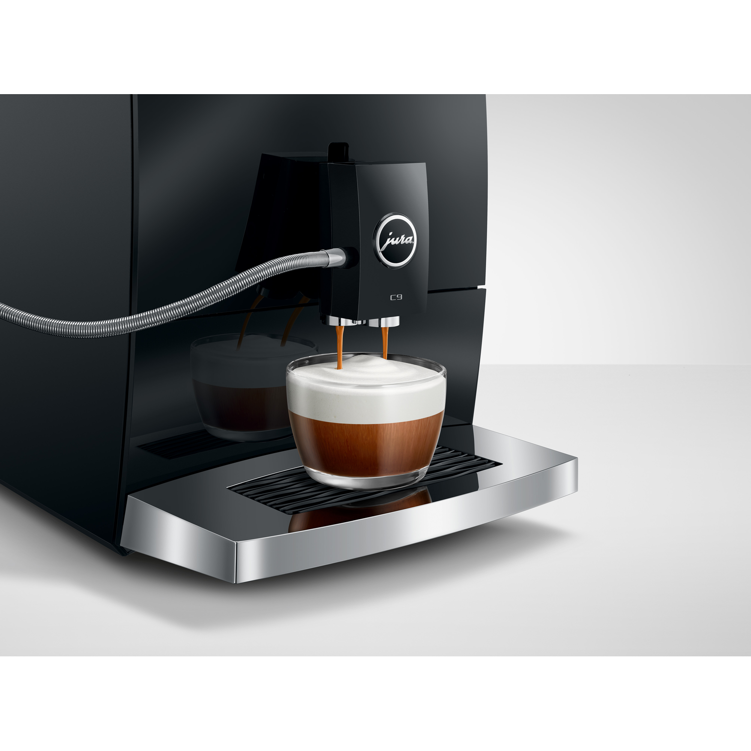 Black Jura C9 coffee machine making a cappuccino. Coffee streams into the glass, topped with white foam, on a silver tray.
