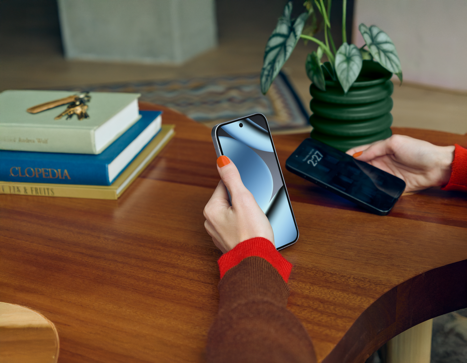 Manos sosteniendo dos teléfonos inteligentes sobre una mesa de madera. Libros, una planta verde y llaves también están en la mesa. La hora en un teléfono es 2:27.