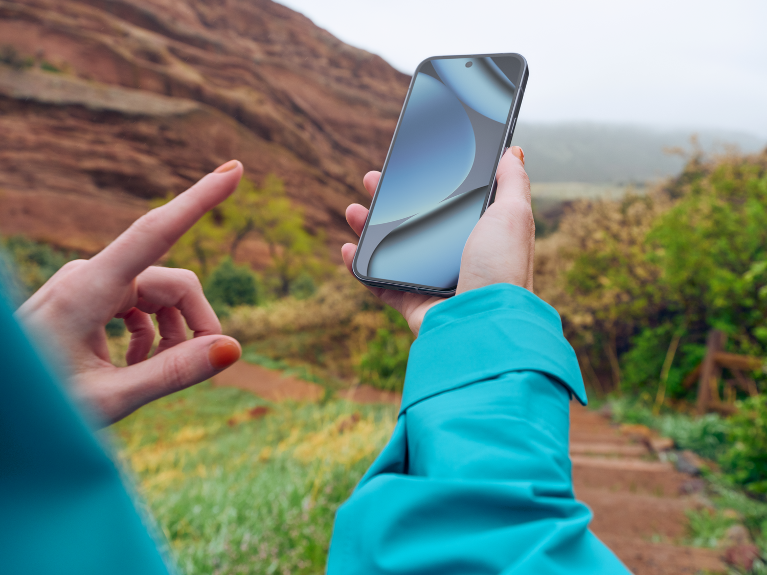 Una persona sostiene un smartphone, señalando con otra mano. El fondo muestra una montaña y vegetación verde.