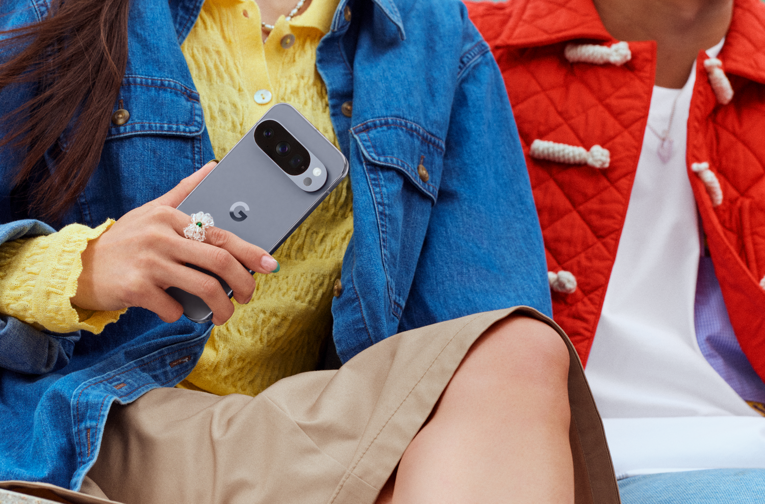 Persona sosteniendo un teléfono gris con cámara. Lleva un suéter amarillo y una chaqueta vaquera, otra persona una chaqueta roja.