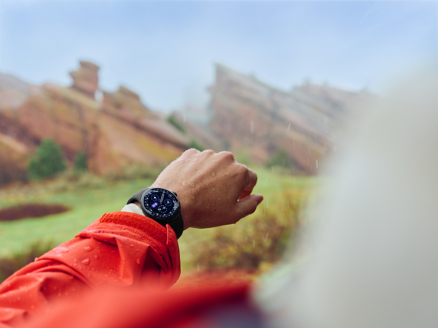 Muñeca con reloj inteligente, chaqueta roja, fondo montañoso y lluvia.