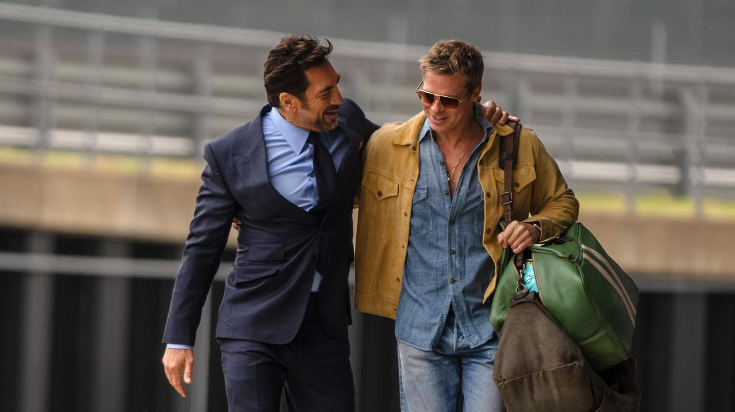 Zwei Männer gehen und lächeln. Einer trägt einen blauen Anzug. Der andere trägt ein Jeanshemd und eine gelbe Jacke und hält eine grüne Tasche.