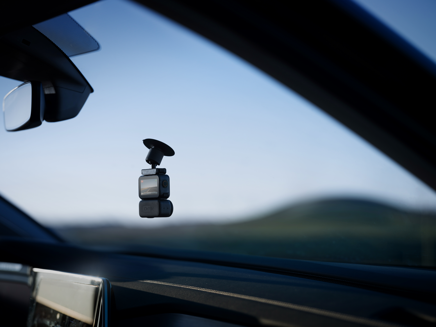 Una cámara de tablero montada en el parabrisas de un coche. El espejo retrovisor es visible. El fondo muestra un paisaje borroso.