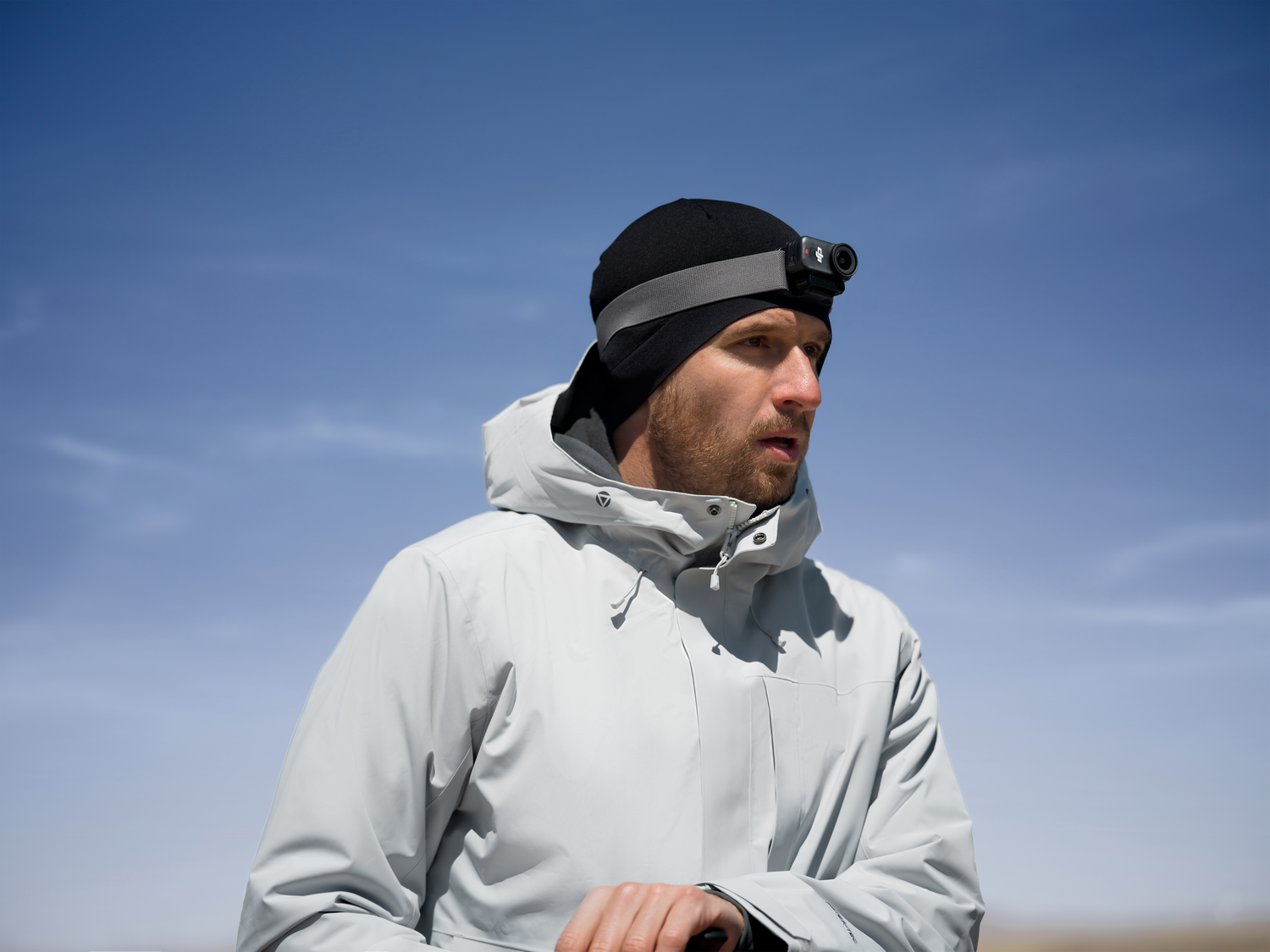 Hombre con gorro negro, lámpara frontal y chaqueta gris contra cielo azul. Mira hacia un lado.