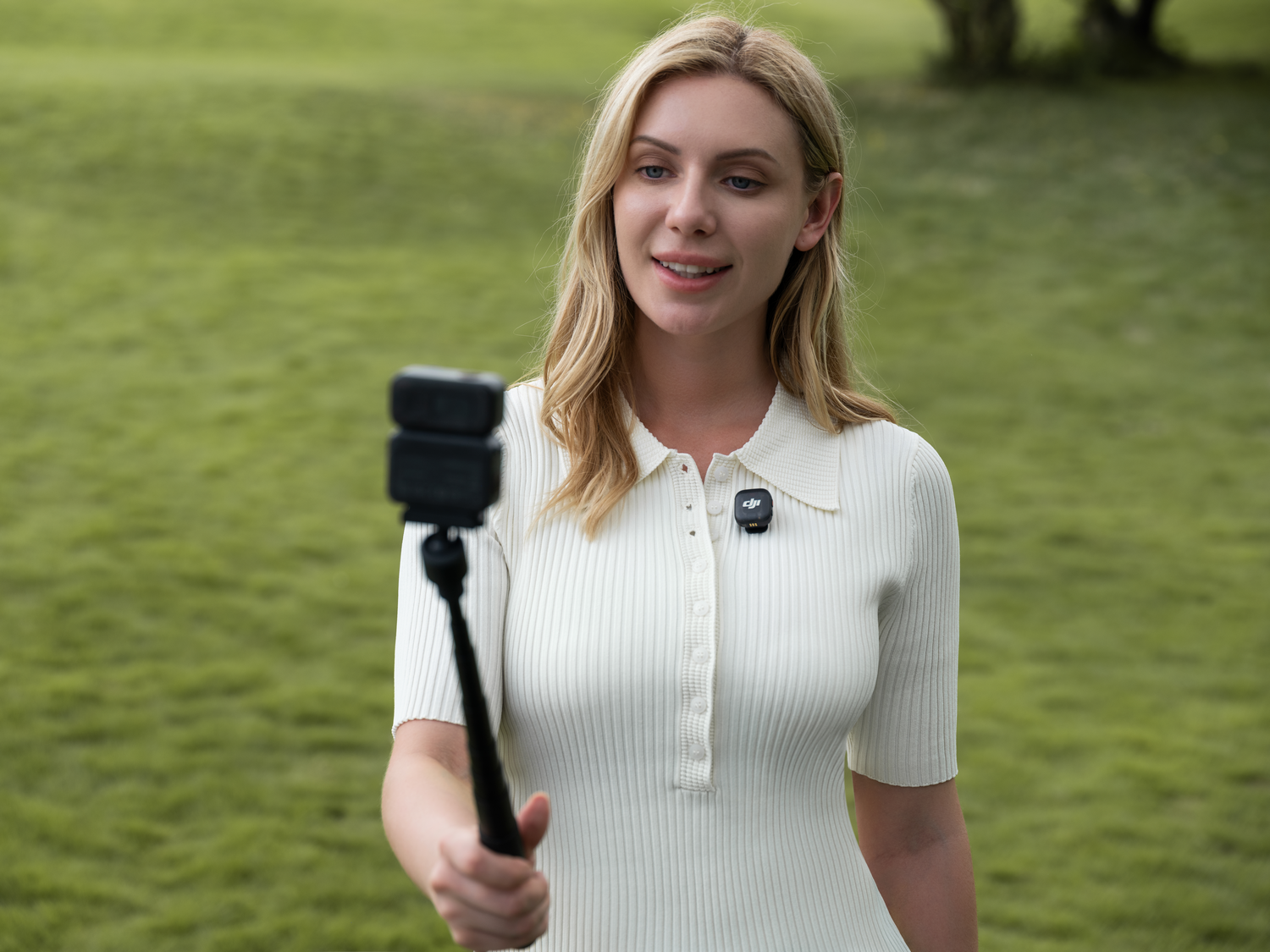 Mujer en vestido blanco con un palo selfie. Sonríe a la cámara con hierba verde de fondo.