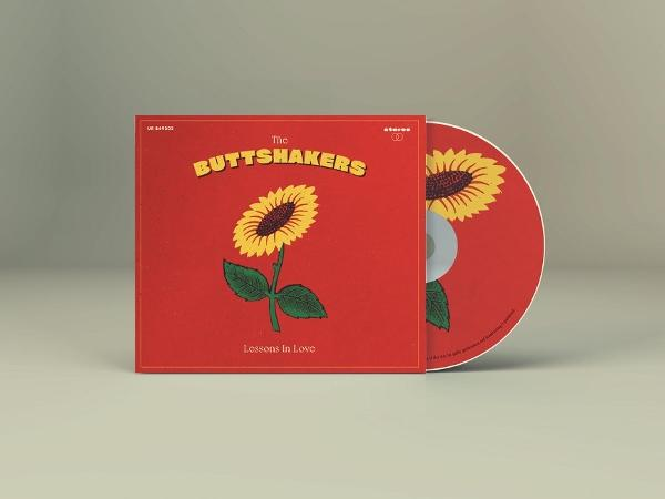 Eine CD-Hülle mit einem Sonnenblumenmuster auf rotem Grund und die CD daneben. Auf der Hülle steht 'The Buttshakers'.