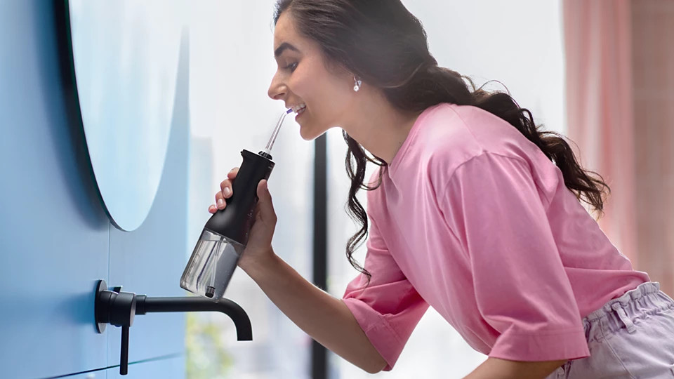 Kobieta używa czarnego irygatora do zębów. Ma na sobie różową koszulę i stoi obok niebieskiej ściany z okrągłym lustrem.