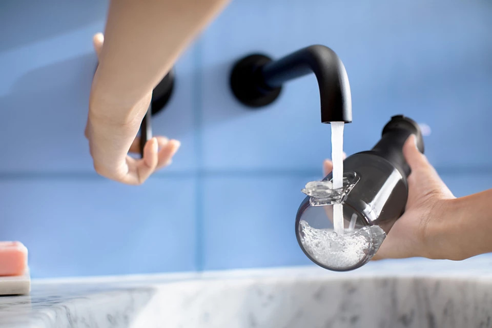 Osoba napełnia czarną butelkę z wodą wodą z czarnej baterii. Tło to niebieska ściana z płytek.