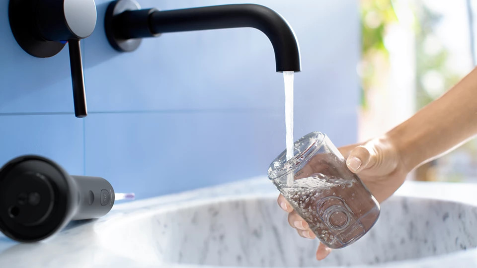 Woda wlewa się z czarnej kranówki do szklanki trzymanej w dłoni. Czarny irygator do jamy ustnej leży na zlewie.