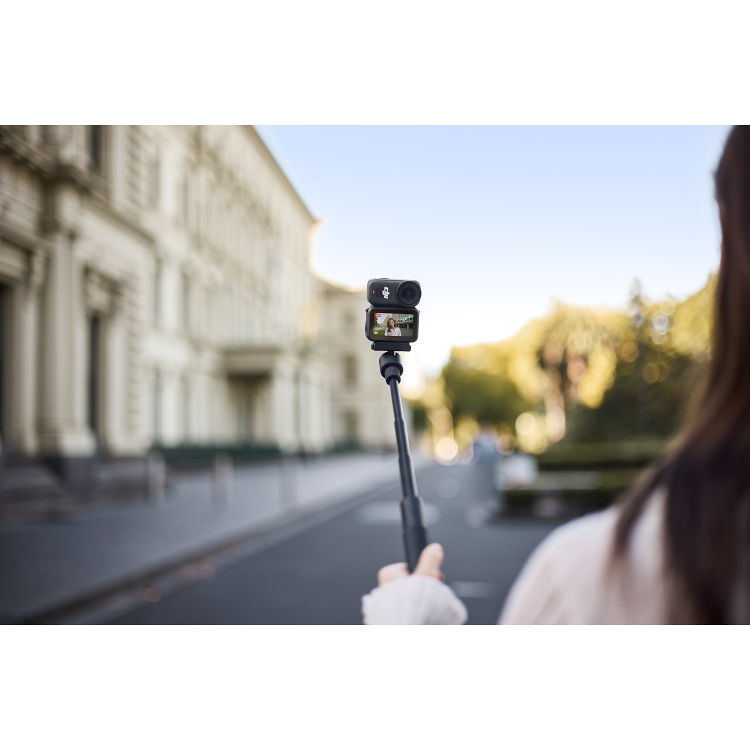 Een vrouw houdt een camera op een selfiestick en filmt een straat met gebouwen. De camera toont een persoon op het scherm.