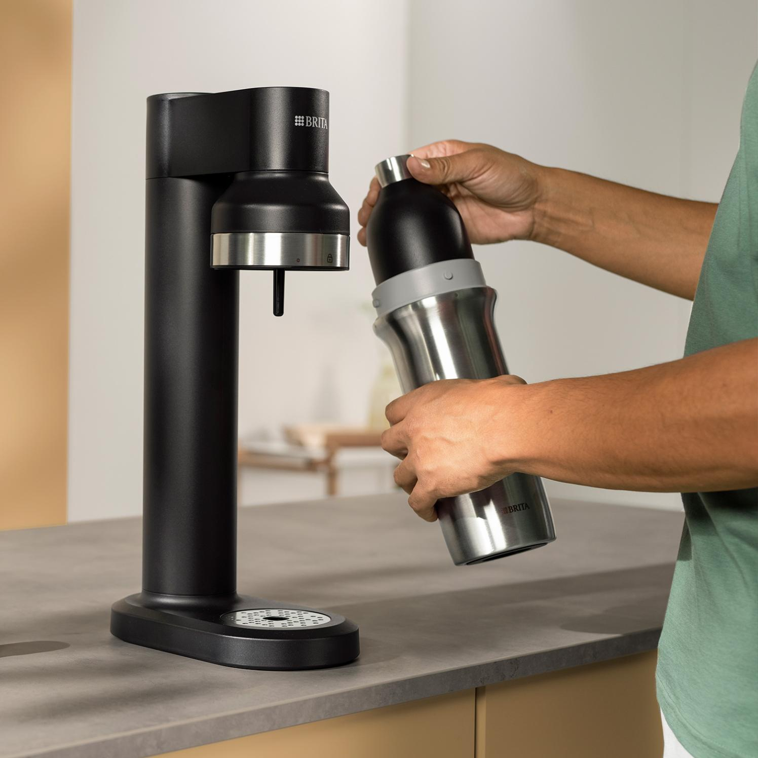 Osoba trzyma butelkę z wodą w pobliżu czarnej maszyny do karbonizacji wody. Maszyna i butelka mają logo Brita.