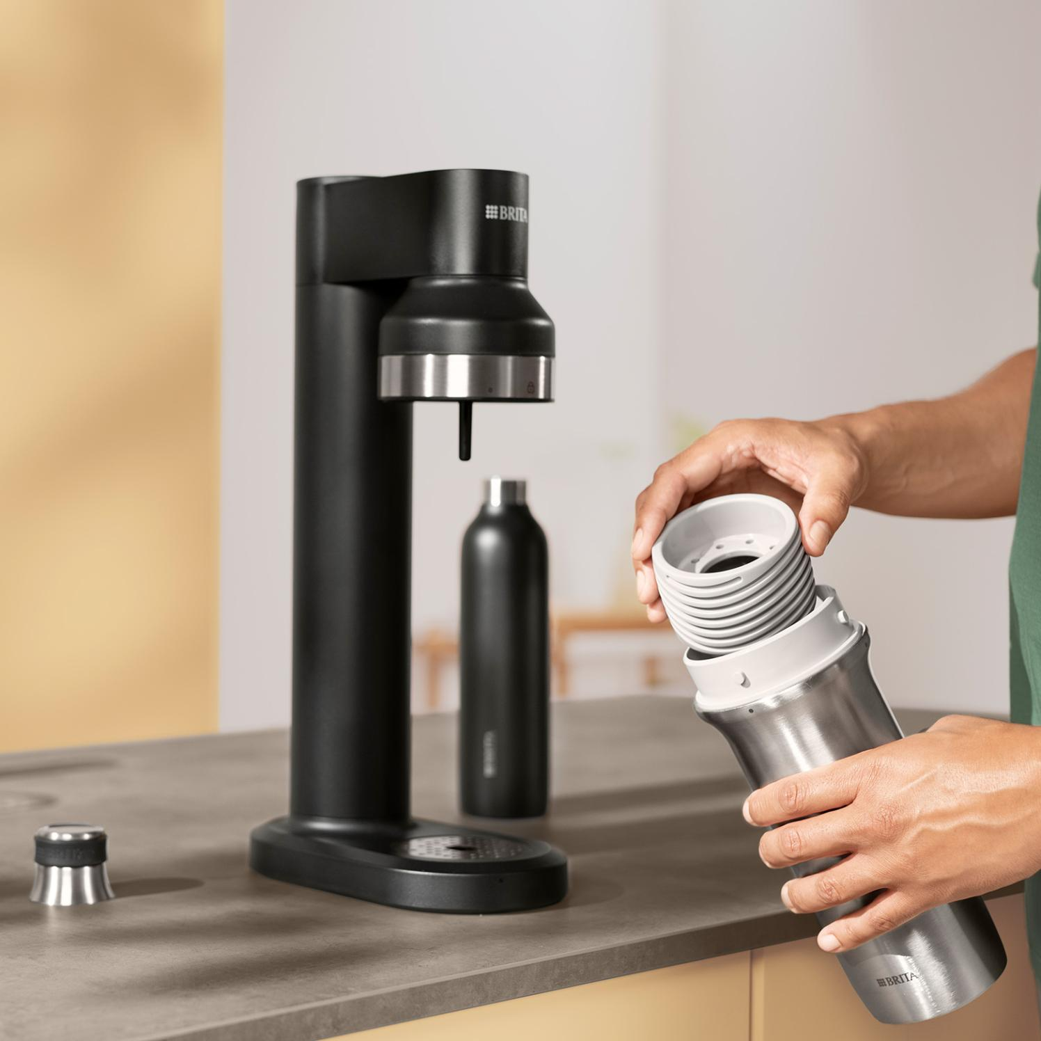 Osoba montuje saturator. Na blacie znajduje się czarna maszyna, srebrna butelka i części. Osoba trzyma część butelki.