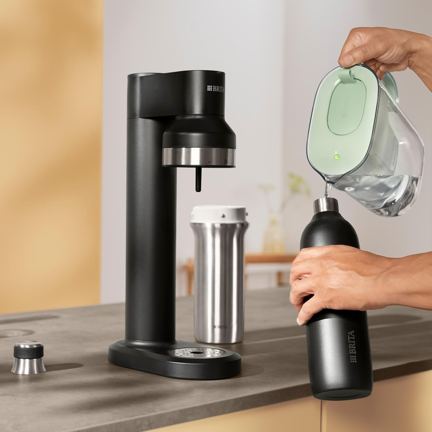 Osoba wlewa wodę z zielonego dzbanka Brita do czarnej butelki Brita. Na blacie znajduje się czarny dozownik wody i srebrna butelka.