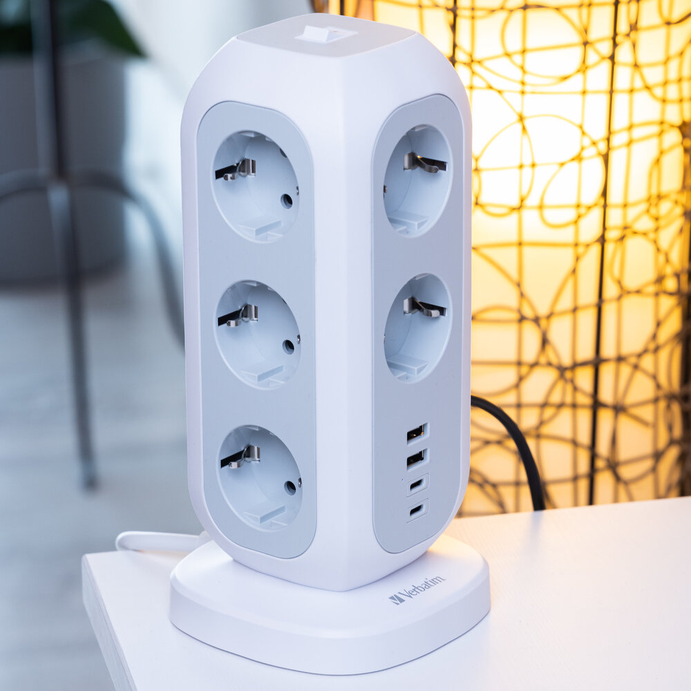 Biały przedłużacz Verbatim z wieloma gniazdami i portami USB stoi na białym stole. W tle świeci lampa.