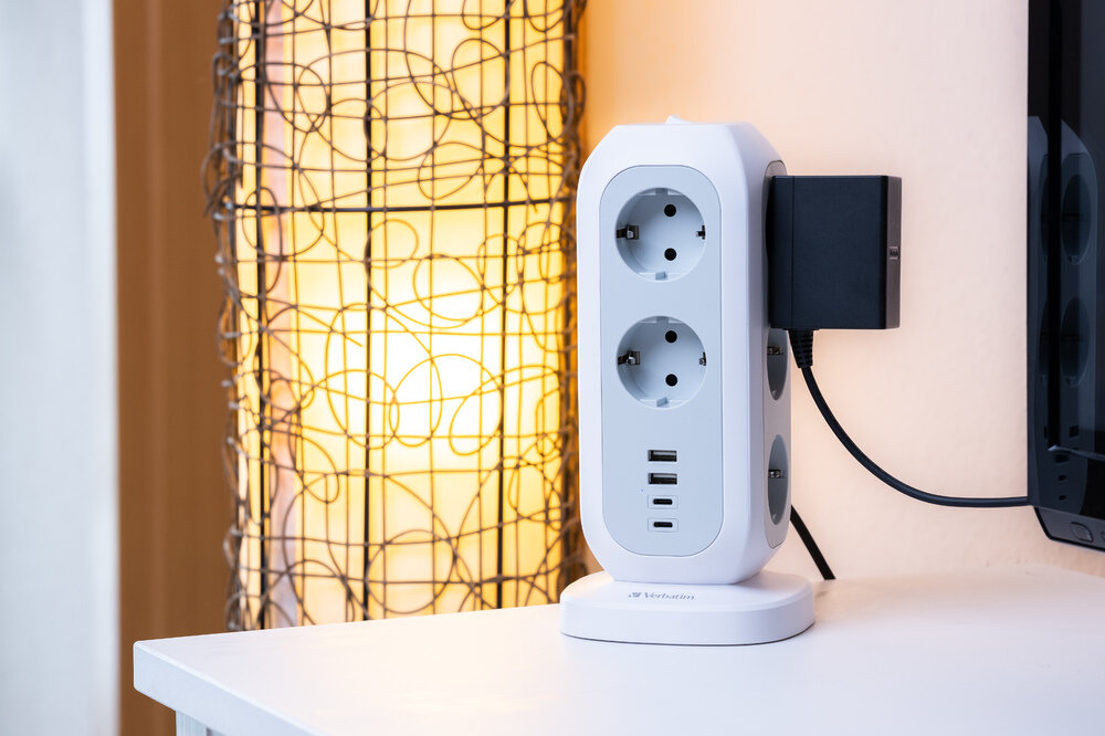 Biała listwa zasilająca z wieloma gniazdami i portami USB. Podłączony czarny adapter. Lampa w tle.