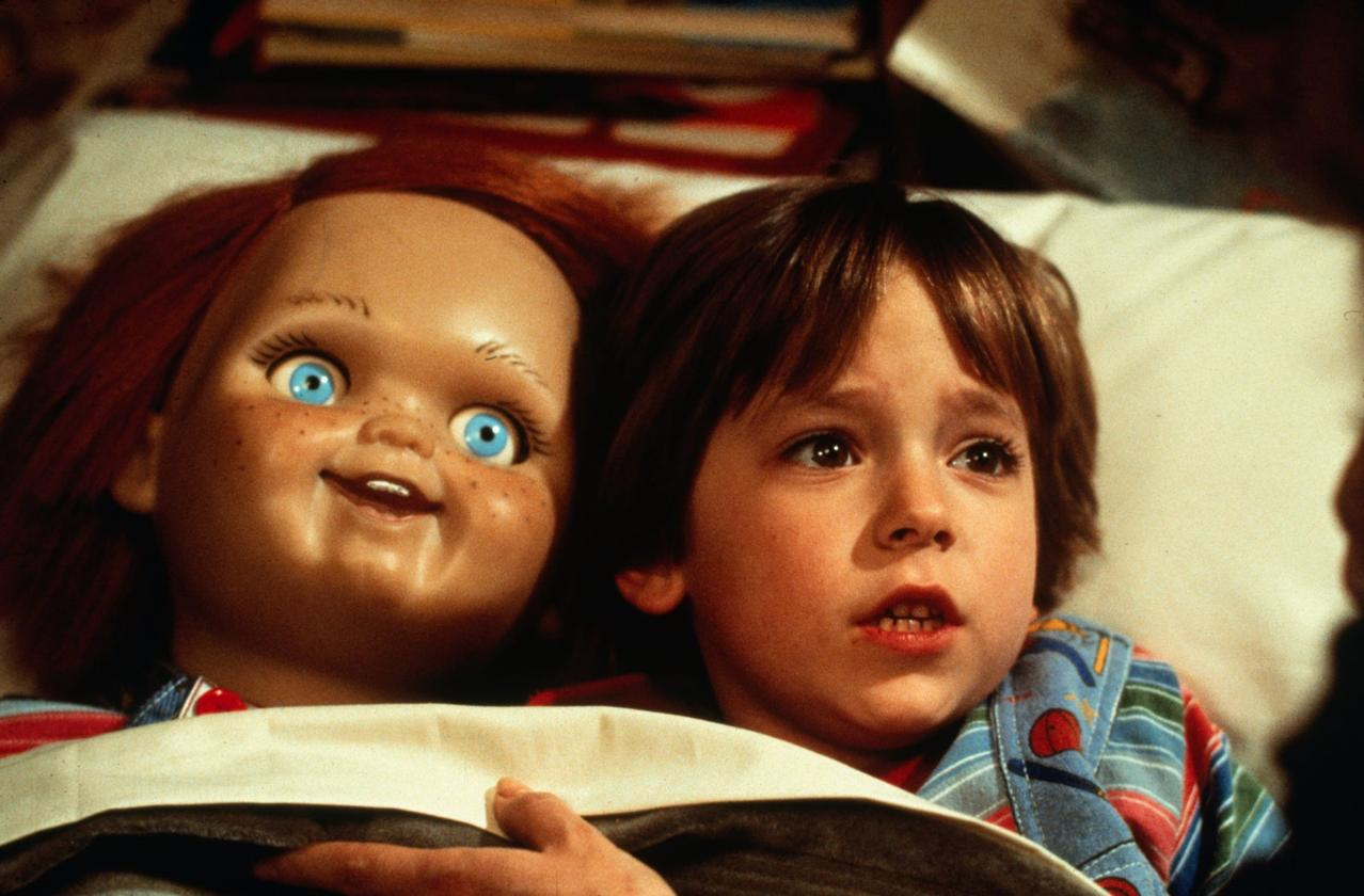 Eine Puppe mit roten Haaren und blauen Augen blickt einen Jungen mit braunen Haaren an. Beide sind in einem Bett. Der Junge sieht verängstigt aus.