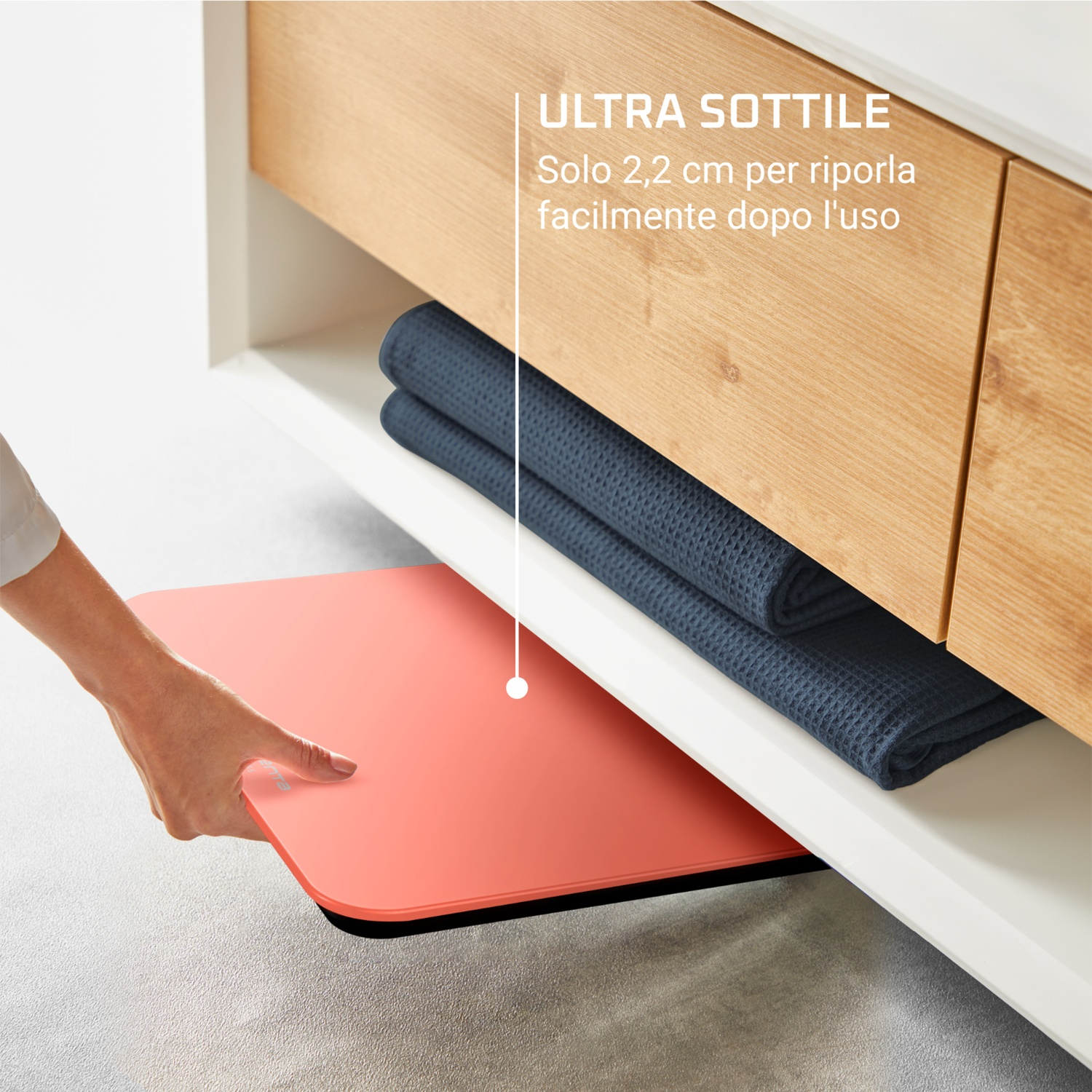 Una mano tira fuori un oggetto rettangolare, sottile, color corallo da sotto uno scaffale con asciugamani blu piegati. Lo scaffale è in un mobile di legno.