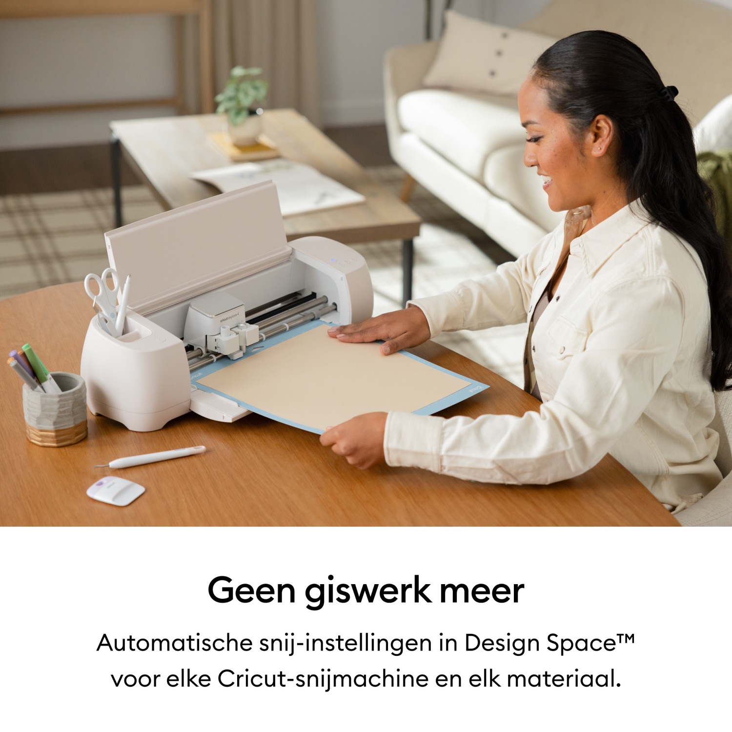 A woman smiles, placing paper into a white cutting machine. Scissors and pens sit nearby on a wooden table, with a couch visible in the background.