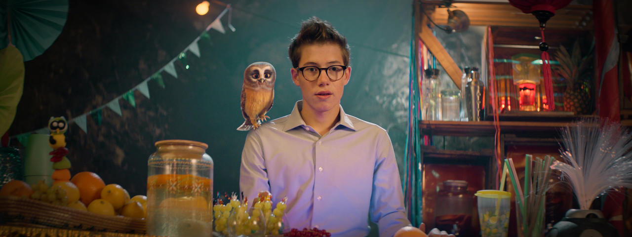 Ein junger Mann mit Brille und einer Eule auf der Schulter steht hinter einer Bar mit Obst und Getränken. Der Hintergrund ist eine Party-Szene.