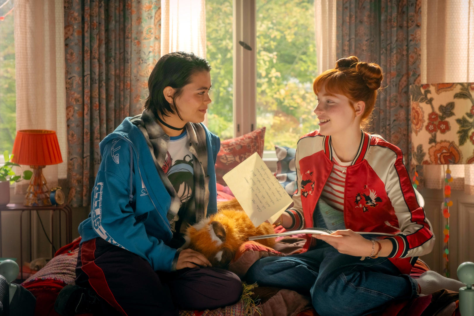 Zwei junge Frauen sitzen auf einem Bett und lesen einen Brief. Eine trägt eine blaue Jacke, die andere eine rote Jacke. Ein Stofftier liegt zwischen ihnen.
