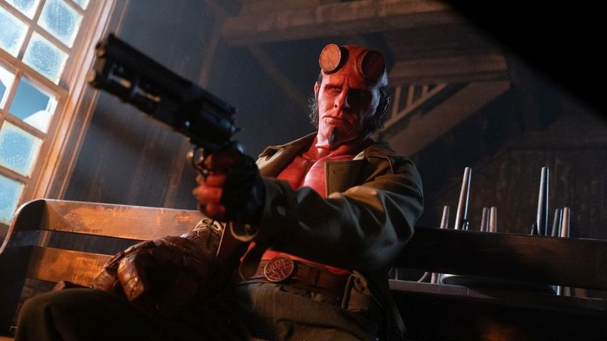 Hellboy, ein rothäutiger Dämon, hält eine Waffe. Er trägt eine Jacke, einen Gürtel. Die Szene spielt drinnen, mit Fenster und Holzbank im Hintergrund.