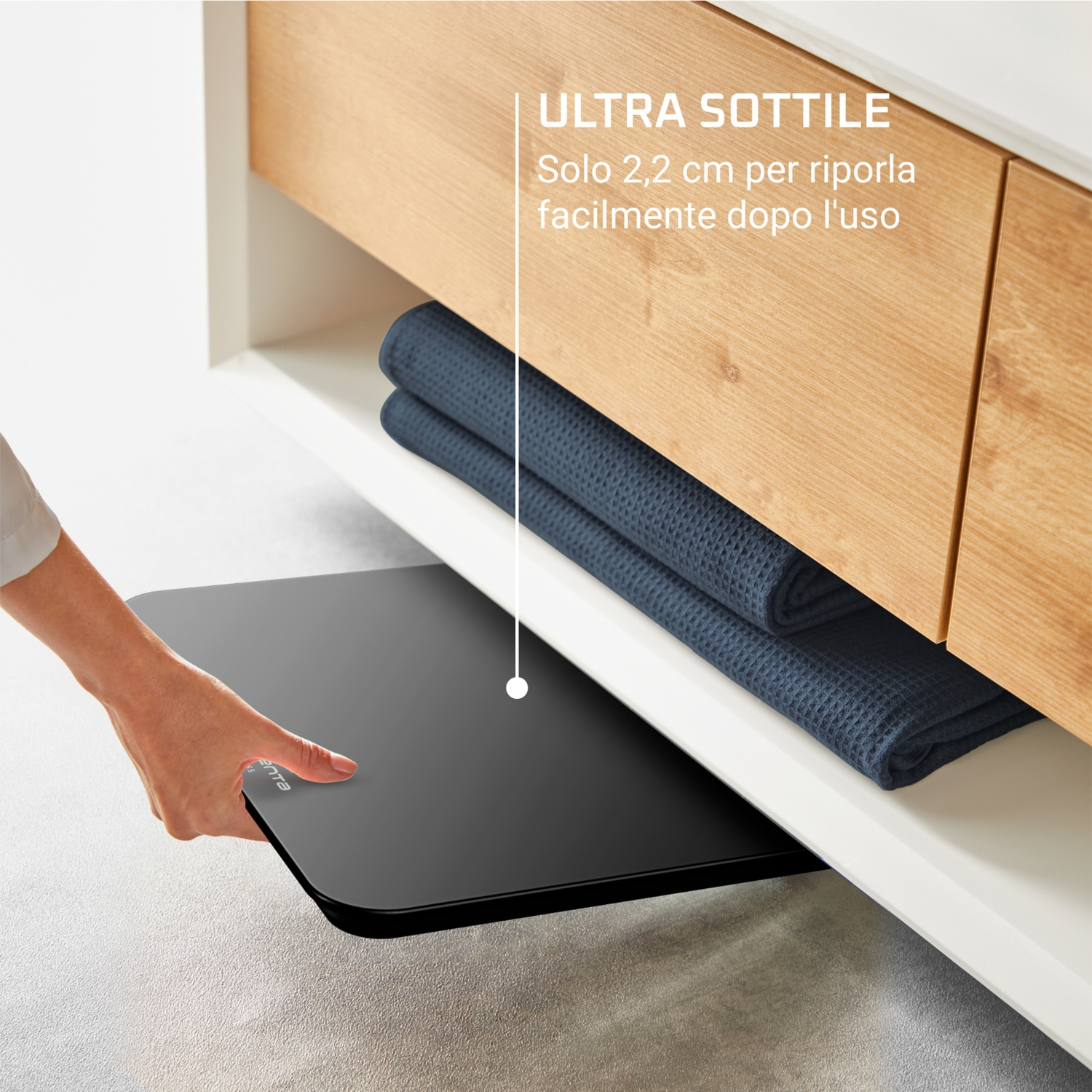 Una mano estrae una bilancia nera da un mobile. Il mobile ha parti in legno e bianche, e all'interno ci sono asciugamani blu.
