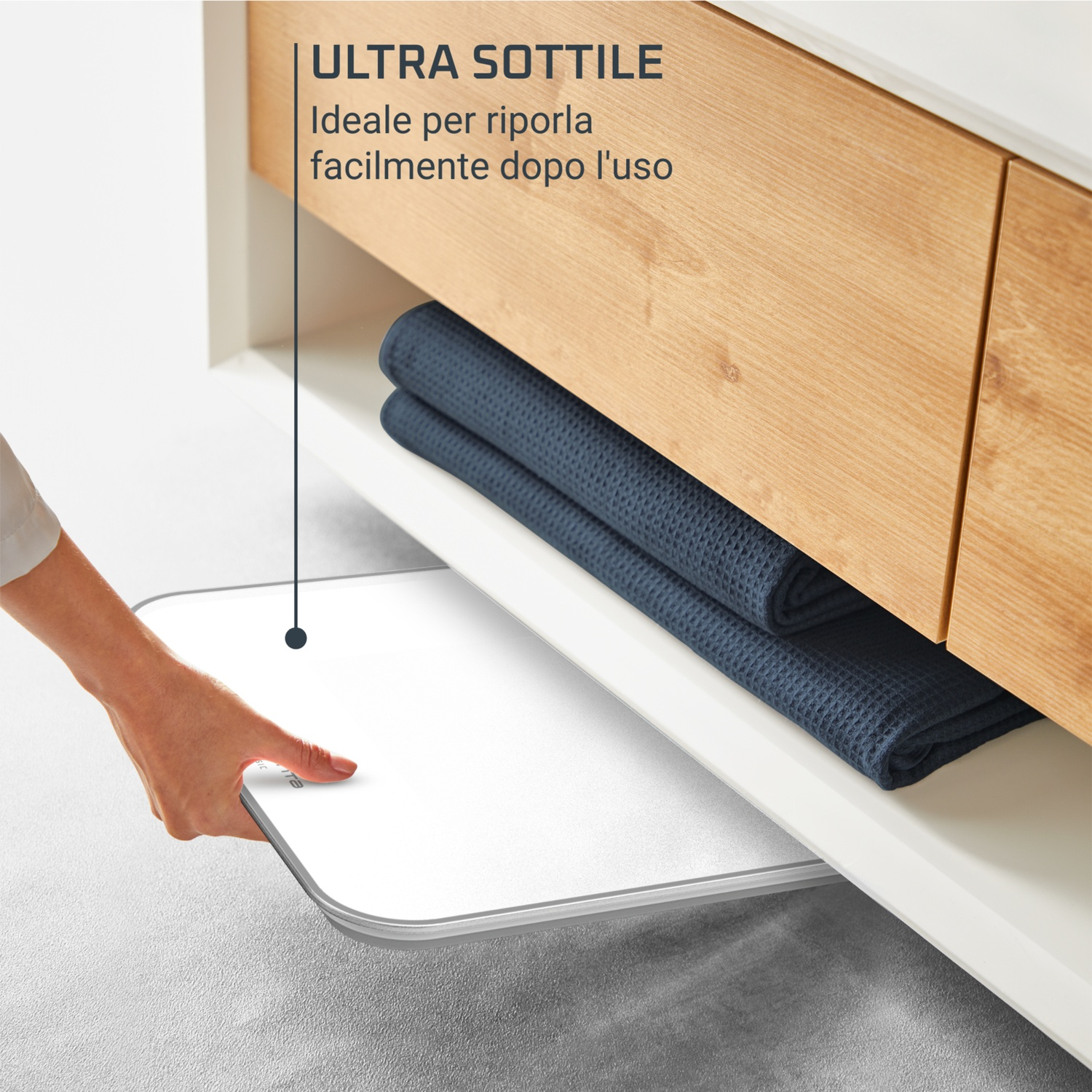 Una mano estrae una bilancia sottile e bianca da sotto un mobile in legno con asciugamani blu. Il testo recita 'Ultra Sottile'.