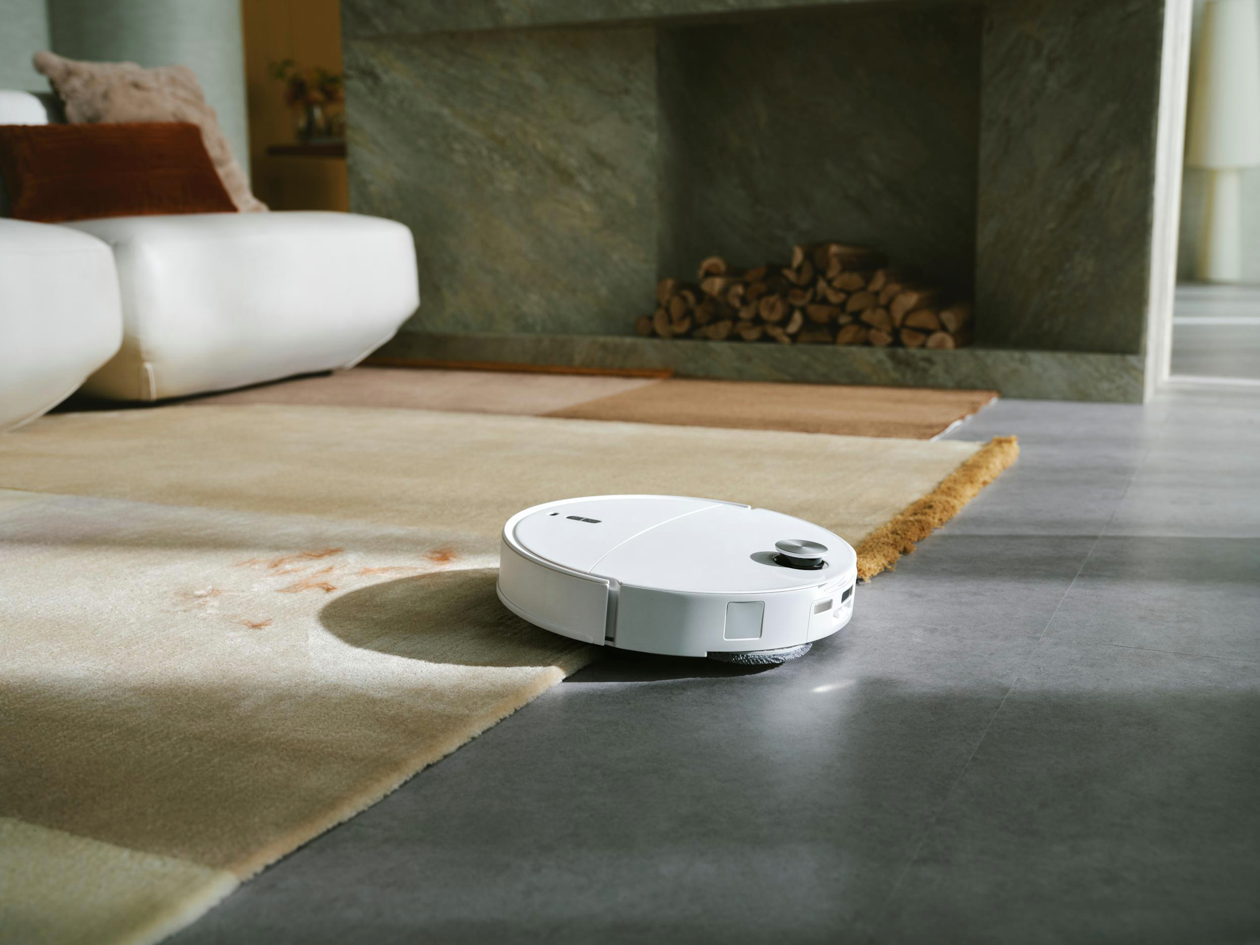 Ein weißer Roboter-Staubsauger auf einem Teppich. Im Hintergrund ist ein Sofa. Kamin mit Holz sichtbar.