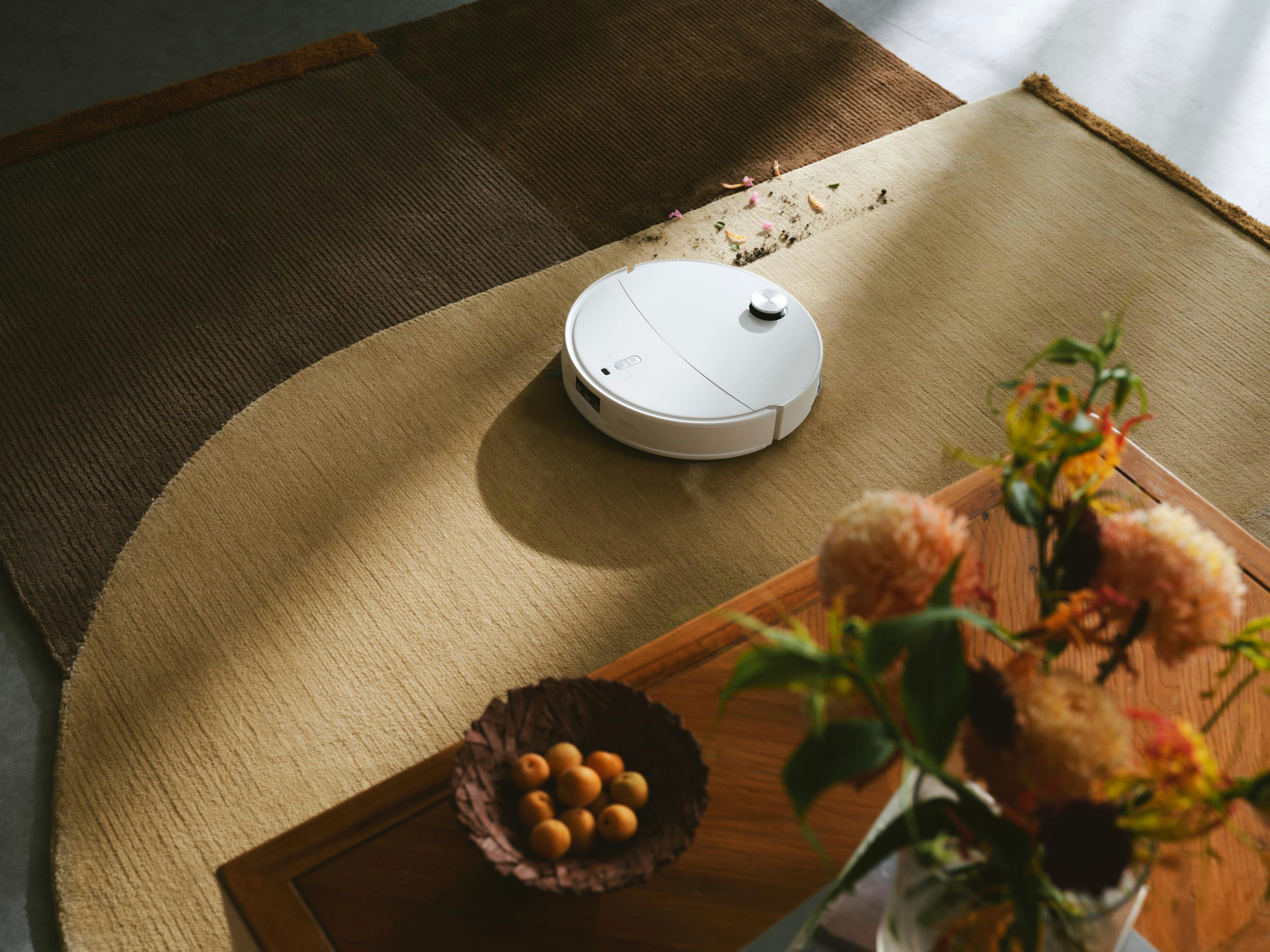 Weißer Roboter-Staubsauger auf gemustertem Teppich, in der Nähe eines Tisches mit Obst und Blumen. Blütenblätter liegen auf dem Teppich.