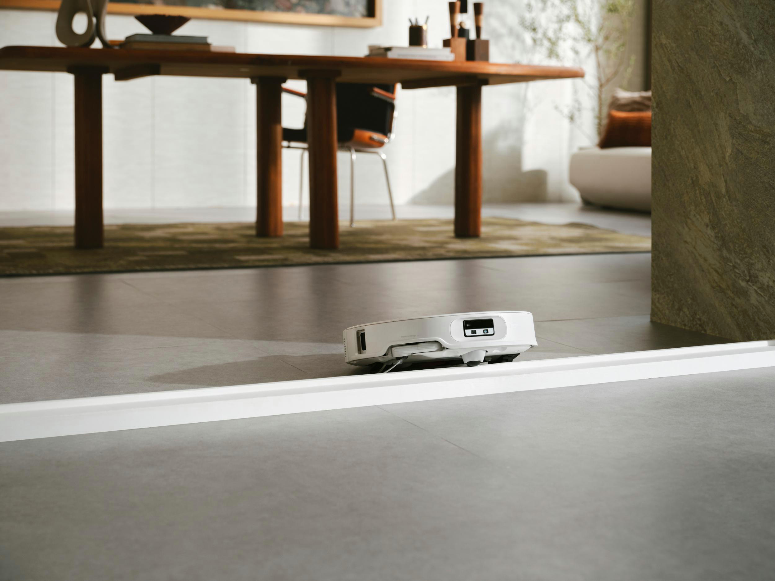 Ein weißer Roboter-Staubsauger auf grauem Boden. Im Hintergrund ein Tisch und ein Stuhl.
