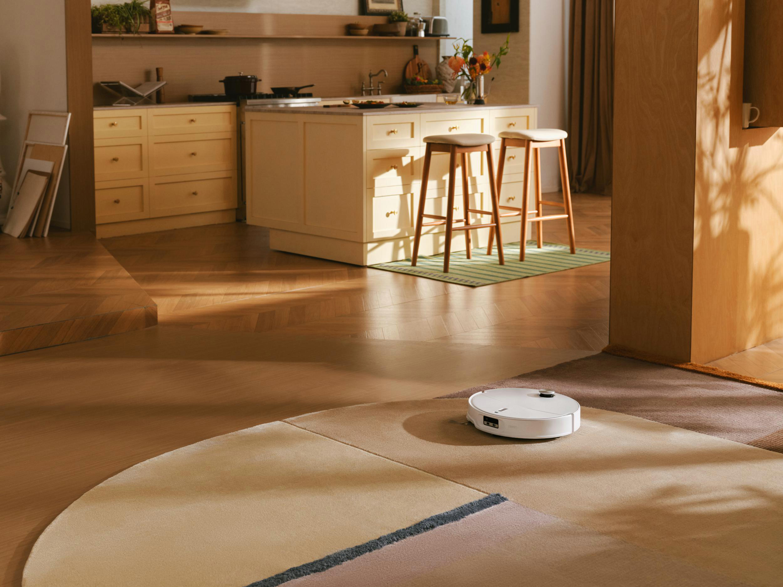 Ein weißer Roboterstaubsauger auf einem Teppich in einer Küche mit einer hellen Insel und zwei Hockern.