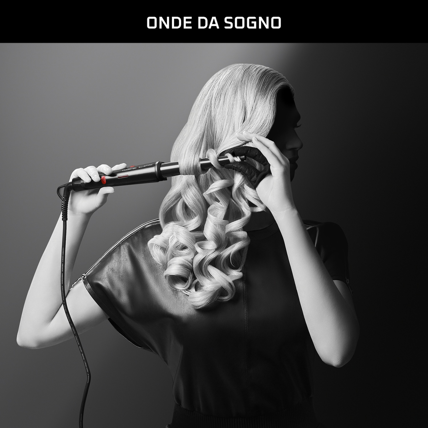 Una donna si arriccia i capelli biondi con un ferro arricciacapelli nero. Indossa un top nero. Lo sfondo è grigio scuro.