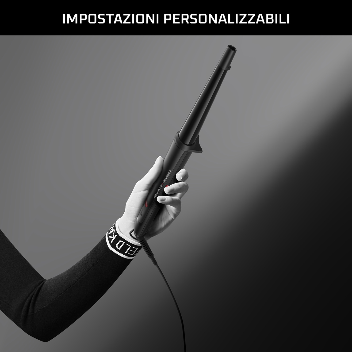 Una mano tiene in mano un arricciacapelli nero. Il braccio è nero e il polso ha un testo bianco. Lo sfondo è grigio.