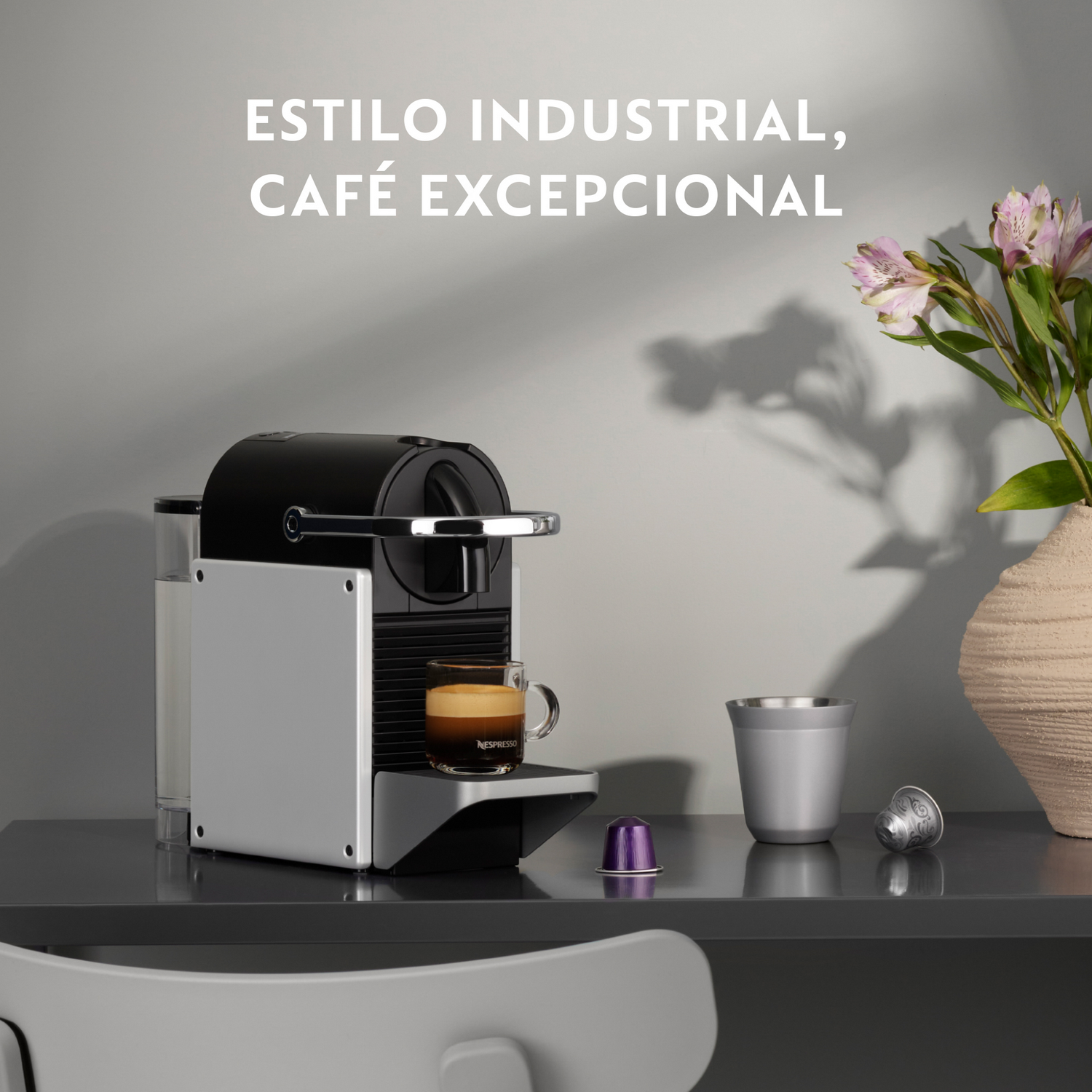 Una cafetera con taza de café, vaso metálico, cápsulas y un jarrón de flores sobre una mesa. El texto dice: Estilo industrial, café excepcional.